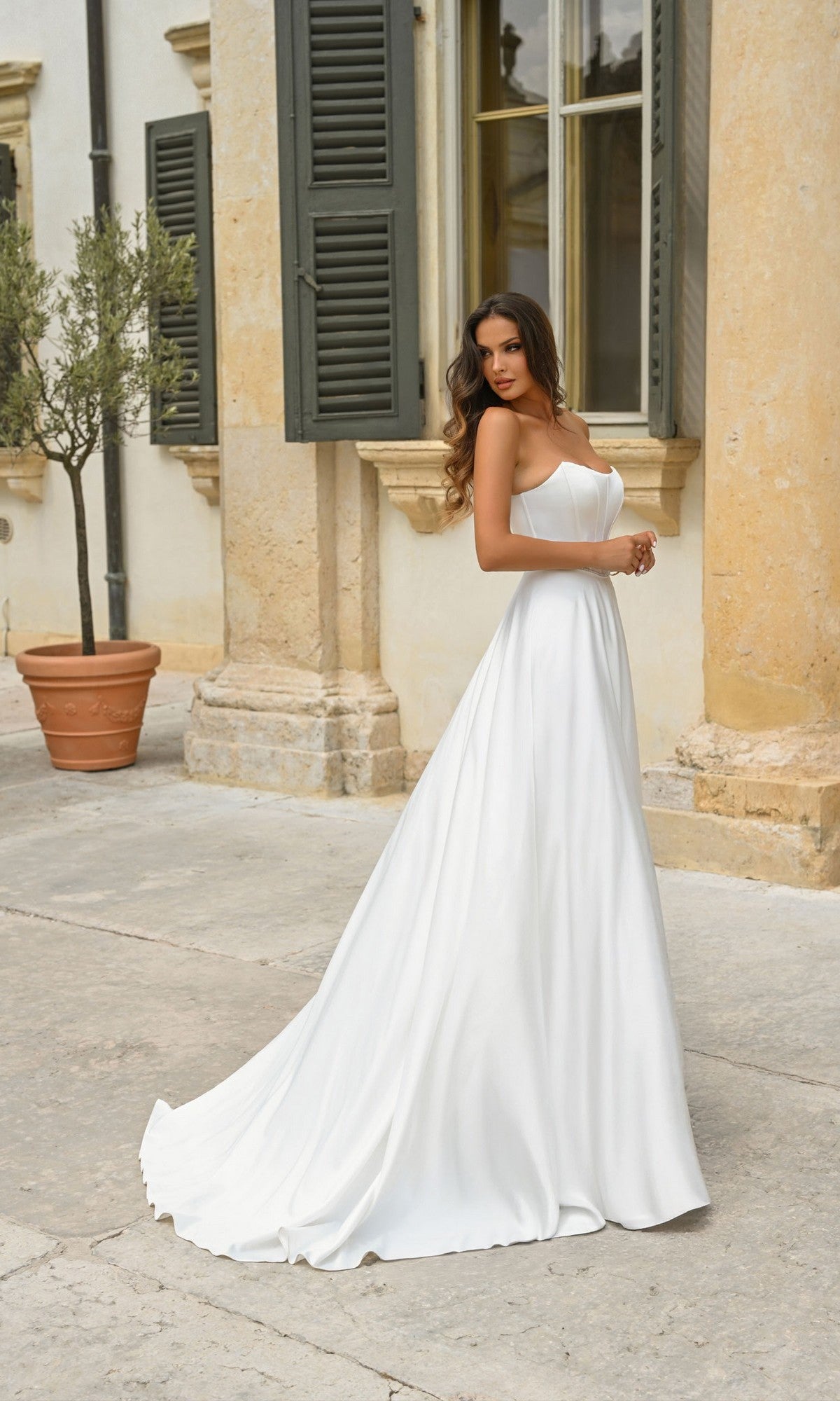 A woman wearing the Alyce 7110 Long White Dress, featuring a fitted silhouette and long train, stands outside a building with large windows and stone columns.